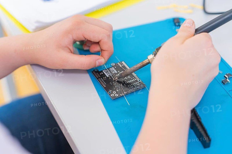 A person solders a circuit board with a soldering iron, surrounded by electronic parts