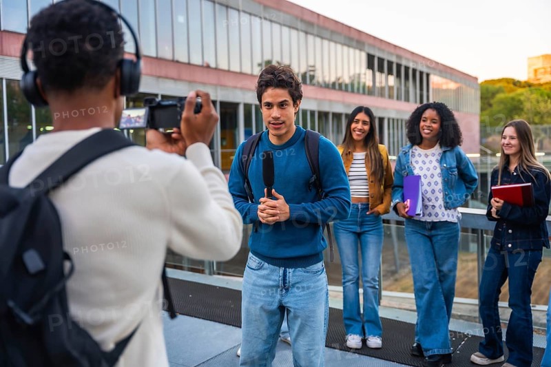 Young man with a microphone speaking while being filmed by another student wearing headphones, with a diverse group of fellow students standing behind on a university campus