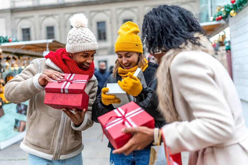Three happy friends opening christmas gifts at a christmas market, enjoying winter holidays together