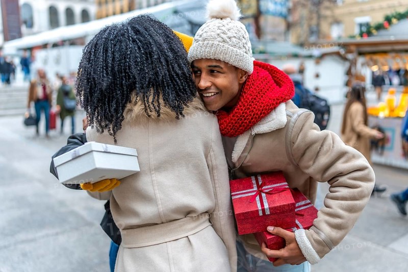Two friends joyfully exchanging gifts at a bustling outdoor christmas market, surrounded by festive decorations and the warmth of holiday spirit