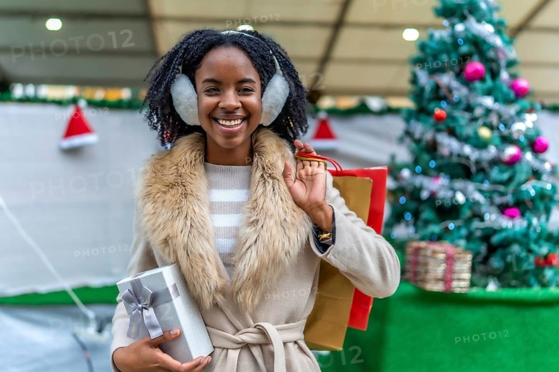 Young black woman wearing earmuffs holding christmas present and shopping bags, smiling in front of christmas tree