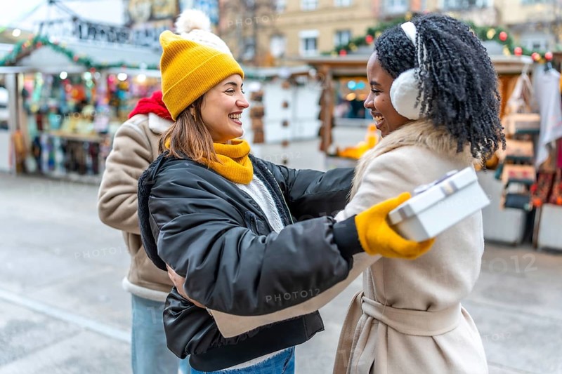 Two happy female friends exchanging christmas presents at a decorated outdoor city market during wintertime