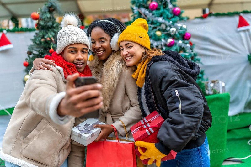 Three happy friends taking a selfie with smartphone at the christmas market, holding gifts and shopping bags