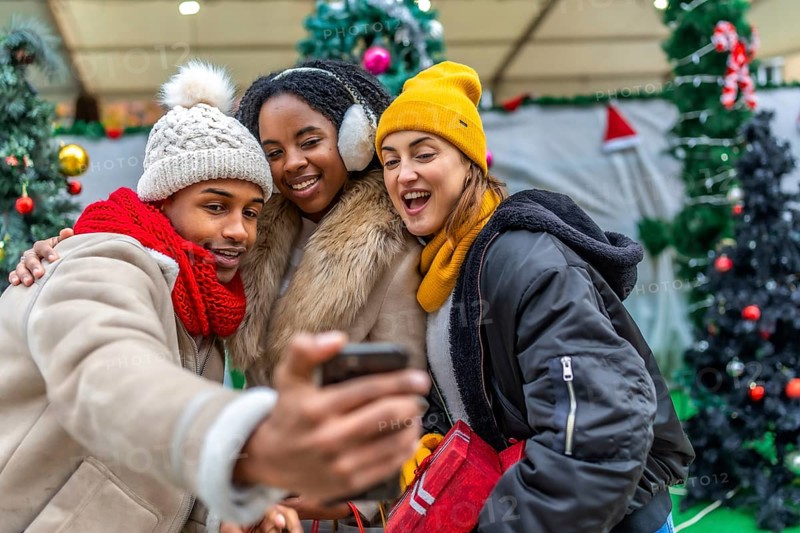 Three cheerful young friends taking a selfie with a smartphone in a city during christmas time, with winter clothes and decorations