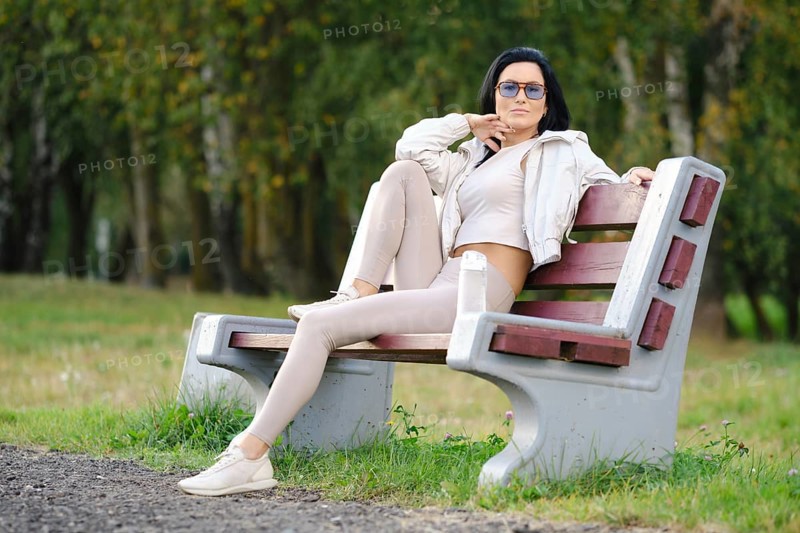 A woman sits on a park bench dressed in light-colored casual clothes, sunglasses on, with a relaxed pose