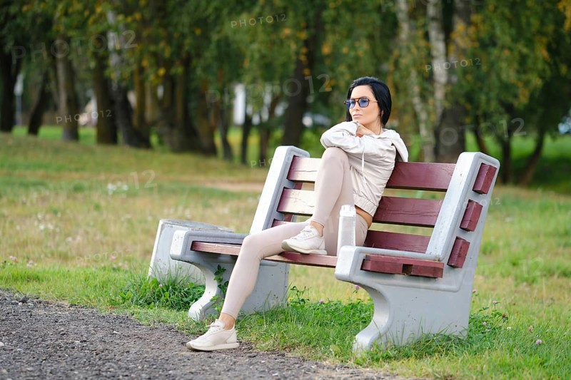 A woman sits on a park bench, wearing leggings and a light jacket. She enjoys the warmth of calm sunny evening and is deep in thought