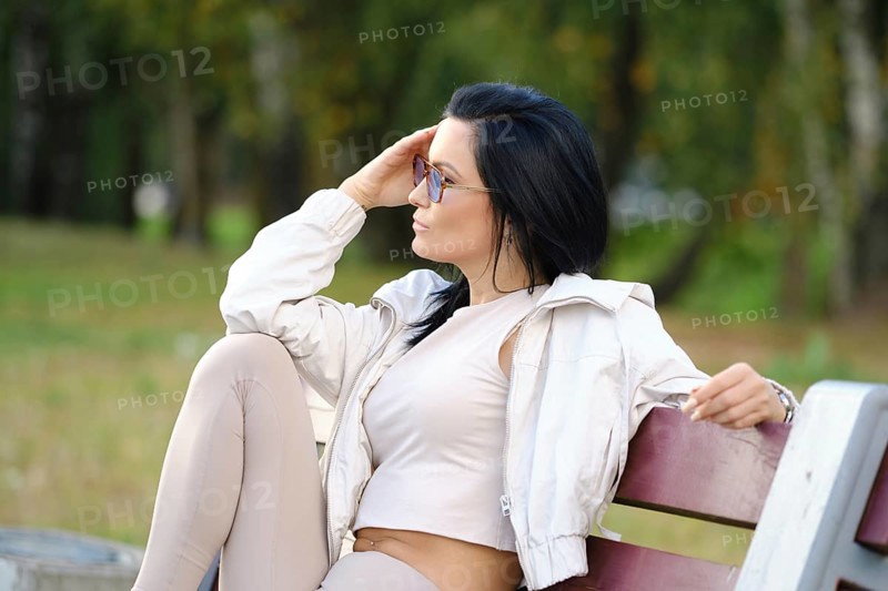A woman with long dark hair sits on a park bench, dressed in comfortable attire. She gazes thoughtfully into the distance, surrounded by lush greenery and soft sunlight filtering through trees
