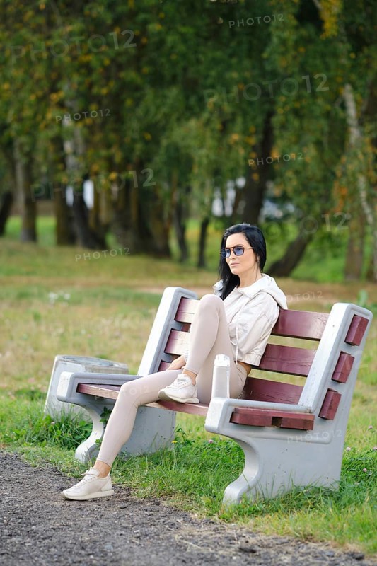 A slender woman sits comfortably on a park bench, wearing glasses and casual attire. Sunlight filters through the trees in the background, creating a serene atmosphere