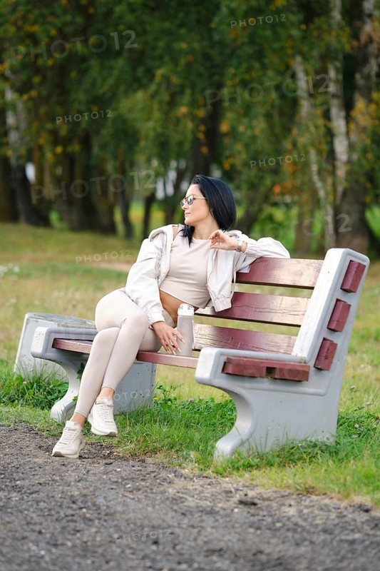 A woman dressed in comfortable casual wear sits on a park bench, gazing thoughtfully into the distance. The serene environment features trees and greenery, creating a peaceful atmosphere