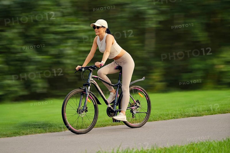 A slender woman in a tank top, cap and leggings quickly rides on a bicycle along the alley in a city park