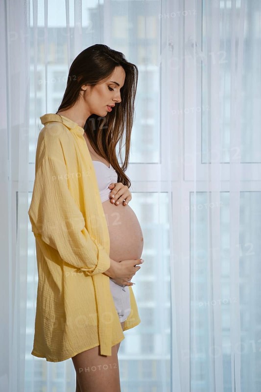 A pregnant woman stands near a window in profile, wearing a yellow shirt and white outfit, lovingly caressing her baby bump. Soft light enters the room, creating a warm and serene atmosphere
