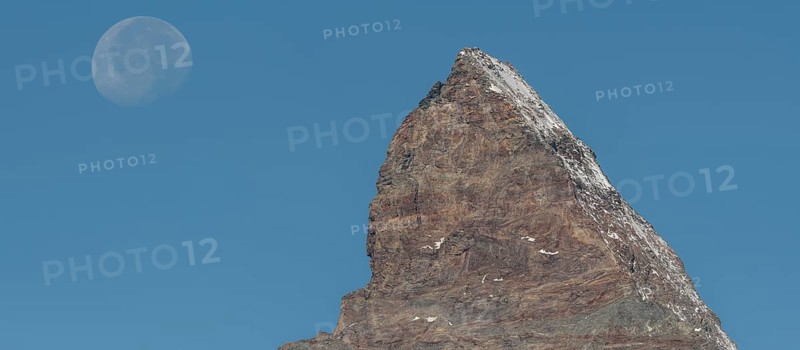 The Matterhorn mountain peak rises steeply against a bright blue sky. The full moon glistens gently above, creating a marvellous view in the serene evening hours. Matterhorn, Zermatt, Valais, Alps, Swiss