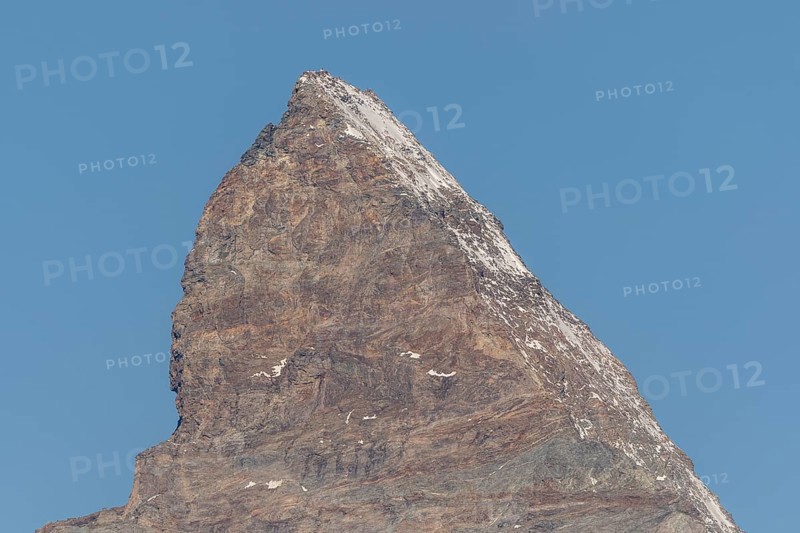 A towering Matterhorn mountain peak stands prominently against the clear blue sky, revealing rocky slopes and a thin blanket of snow on its summit during the bright midday sun. Zermatt, Valais, Alps, Swiss