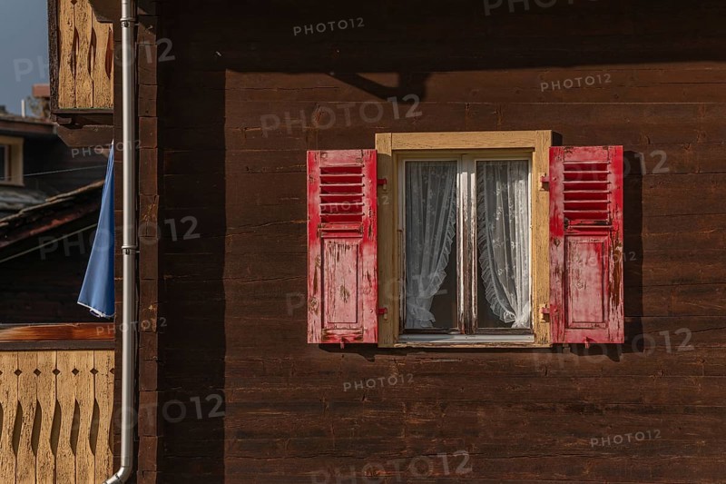 The wooden house stands in a mountain landscape, the windows are decorated with red shutters. Lace curtains add a welcoming touch to this natural atmosphere. Grachen, Viege, Valais, Switzerland