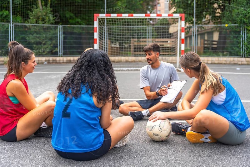 Soccer coach sitting on the ground with clipboard explaining game strategy to female players sitting in a circle around him during a break on the field