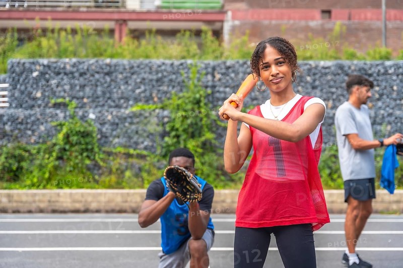 Young woman baseball player holding orange bat ready to hit the ball during a game, with a male teammate ready to catch the ball and another teammate waiting his turn in the background