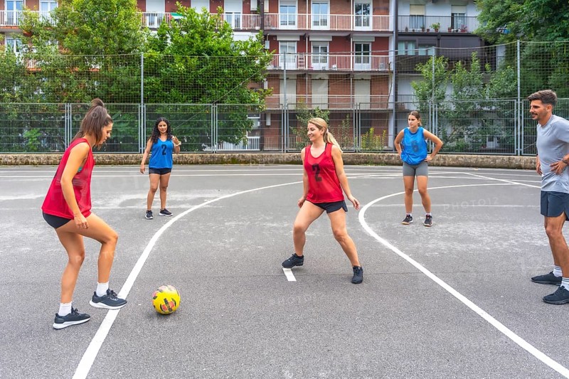 Female soccer players in red and blue jerseys kicking a yellow soccer ball during a friendly outdoor match, highlighting teamwork and athleticism in a neighborhood setting