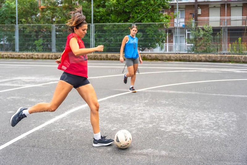 Two women athletes playing soccer on an outdoor court are passing the ball to each other, showcasing their athleticism and teamwork in an urban environment