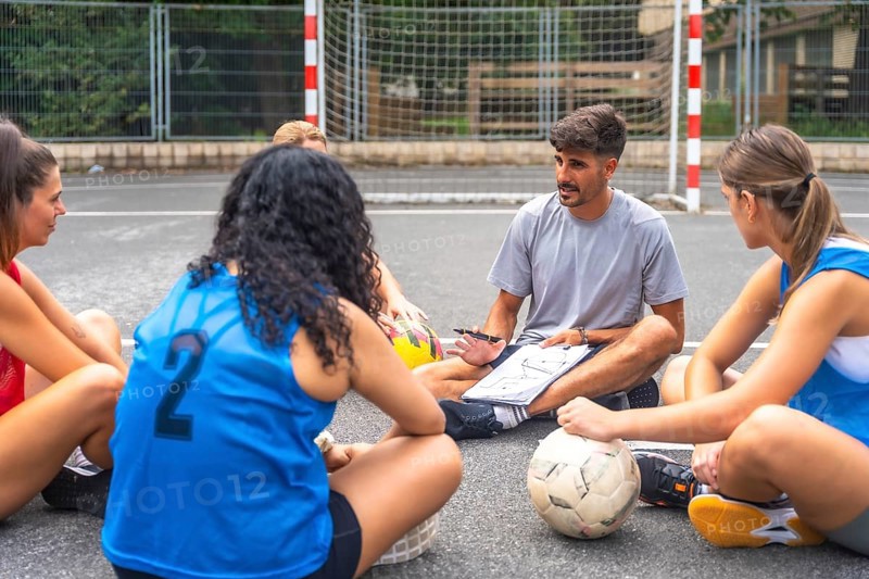 Soccer coach sitting on the ground, using a notebook to explain game strategies to female athletes during an outdoor training session on the court, fostering teamwork and focus