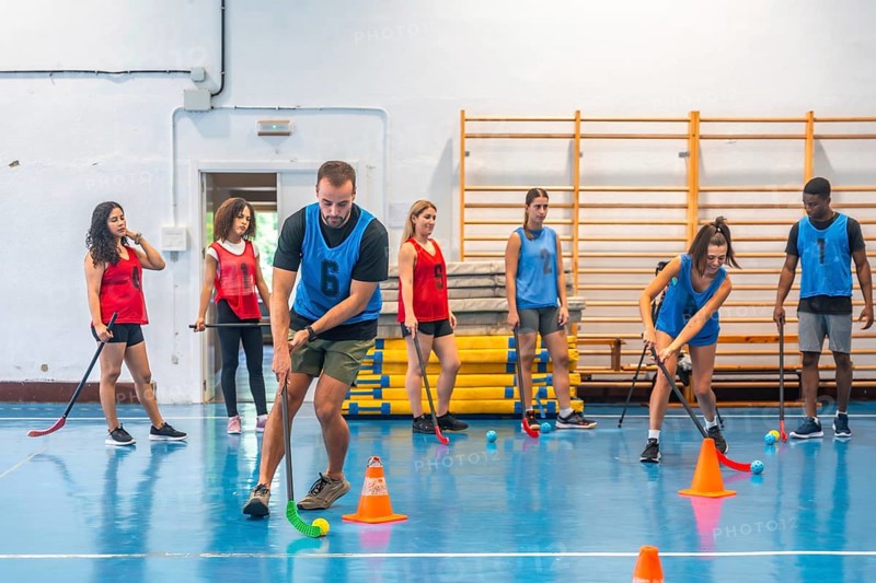 Floorball players practicing drills on court with coach watching, focusing on stick handling skills and teamwork during training session in gym