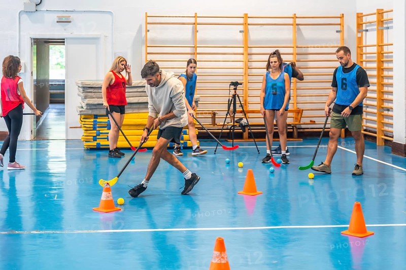 Group of athletes practicing floorball, passing the ball with sticks and maneuvering around cones in a gym, showcasing teamwork and athleticism
