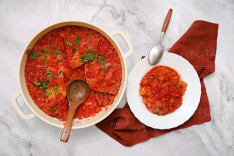 A hearty dish of pappa al pomodoro is in a pot alongside a serving on a white plate. Fresh herbs garnish the rich tomato and bread soup, inviting enjoyment of this traditional Italian comfort food