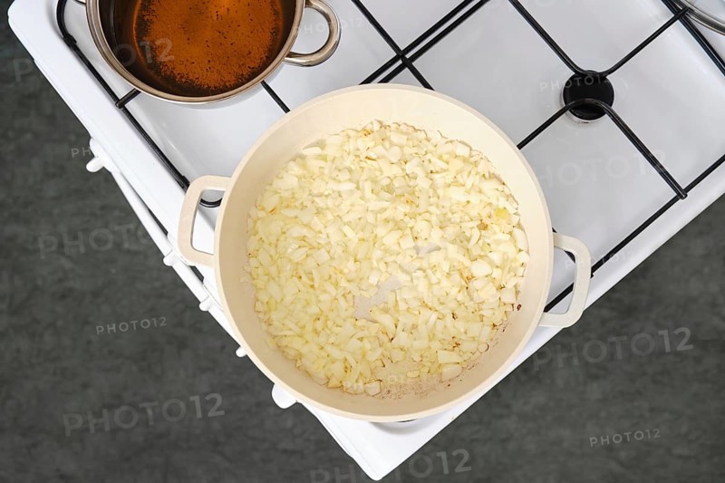 Chopped onions cooking in a white pan over medium heat on a stove. A pot of simmering broth sits close by, releasing steam into the kitchen air, suggesting a flavourful dish in preparation