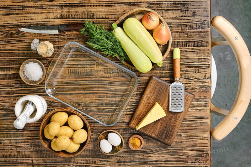 Fresh ingredients for loaded baked potato casserole are displayed on a wooden table. Potatoes, zucchini, onions, and cheese await preparation alongside seasonings and eggs for a delicious dish