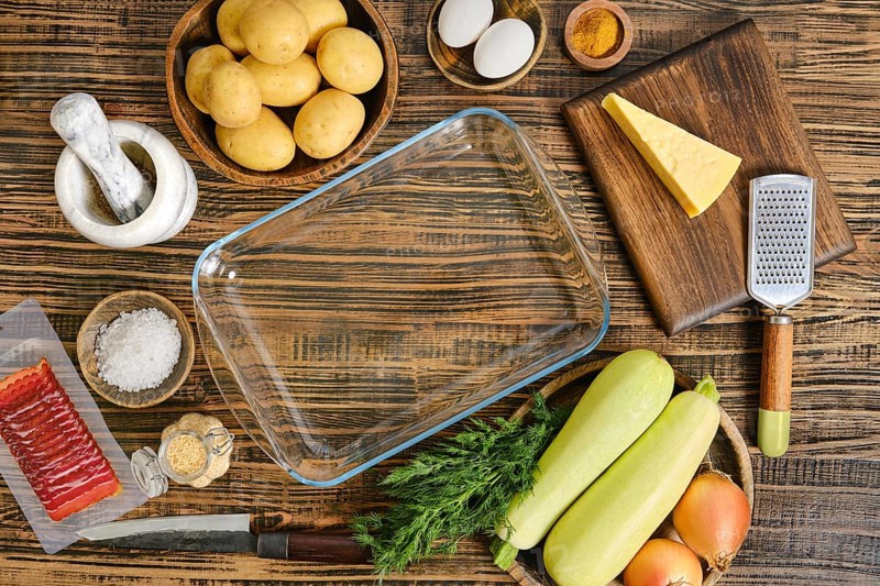 A variety of fresh ingredients are arranged on a wooden table, including potatoes, zucchini, cheese, and eggs. A glass baking dish and kitchen tools are also visible, ready for casserole preparation