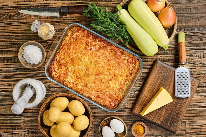 Loaded baked potato casserole sits in a glass dish surrounded by fresh vegetables and spices. Potatoes, cheese, and eggs are prepared alongside zucchini and onions, creating a wholesome meal