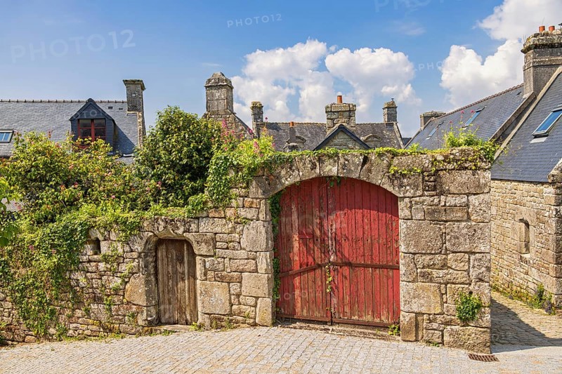 Red wooden doors in an old masonry wall with lush green plants in an old town by a square a sunny summer day, Locronan, Crozon Peninsula, Bretagne, France