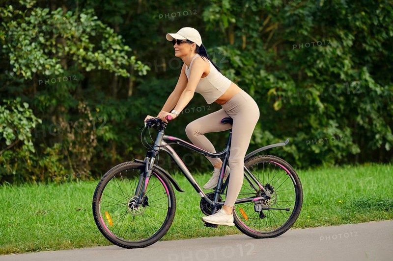 A woman enjoys riding her mountain bike along a serene path in a park. Dressed in fitted activewear, she pedals confidently while surrounded by lush greenery under a bright sun