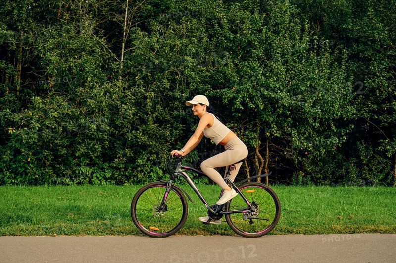 A woman rides a bicycle along a smooth path with vibrant green grass on one side and dense trees lining the other. The sun shines brightly, indicating a pleasant day for outdoor activities