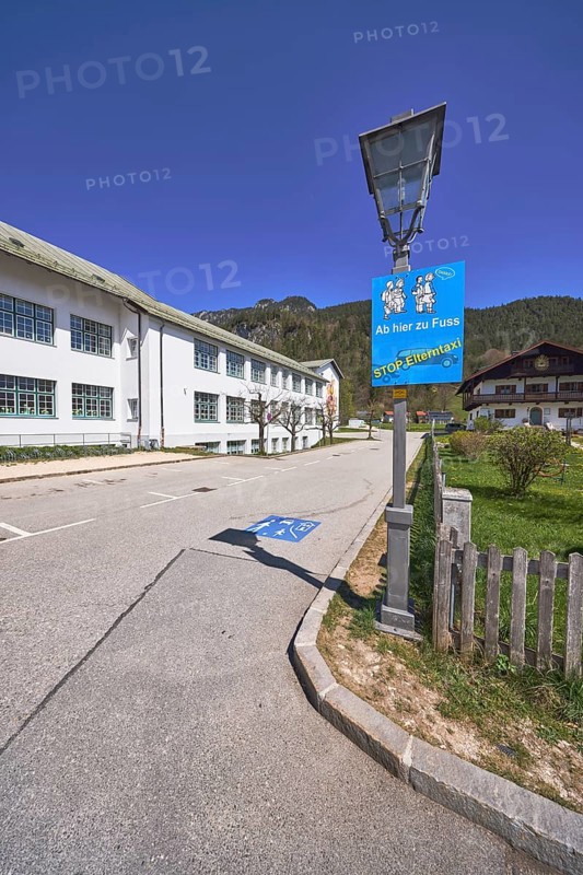 Primary and secondary school Bischofswiesen, sign Stop parent taxi, building, lantern, problem parent taxi, wooden fence, hill, coniferous forest, blue sky, cirrostratus clouds, intersection Göllstraße with Schulstraße, Bischofswiesen, district Berchtesgadener Land, Bavaria, Germany