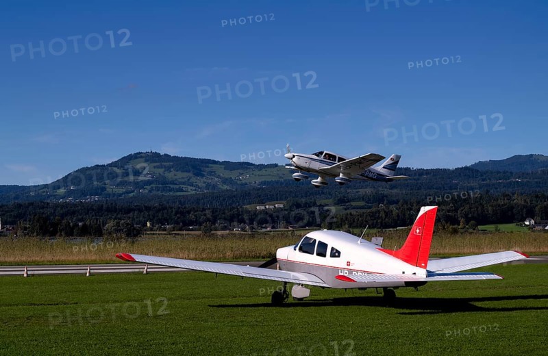 PA28 Archer II 180 HP identification aircraft registration HB-PMG during take-off over Piper Archer II HB-PPY on the ground, Wangen-Lachen airfield, LSPV, Canton Schwyz, Switzerland