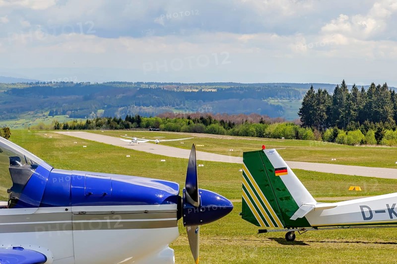 In the background runway and landing strip on it tow launching motorised aircraft towing glider glider taking off, in the foreground left nose tip of parked single-engine propeller aircraft right rudder of motor glider in parking position, Wasserkuppe airfield, Gersfeld, district of Fulda, Hesse, Germany