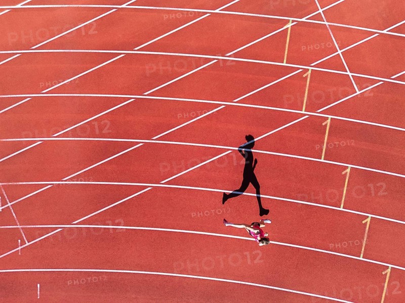 Aerial view of a female individual (runner) on a red athletics track (tartan track) with shadow as a silhouette