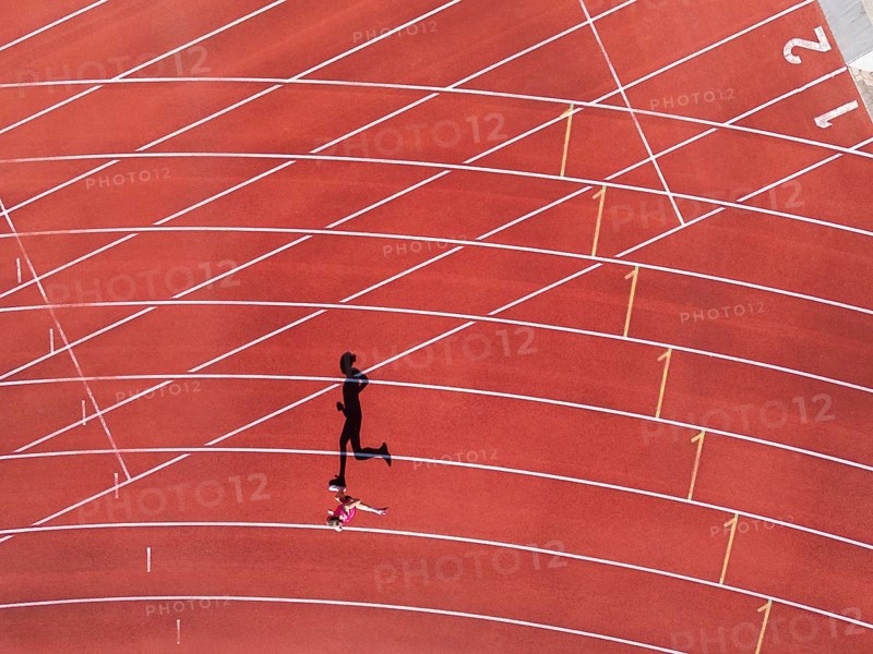 Aerial view of a female individual (runner) on a red athletics track (tartan track) with shadow as a silhouette