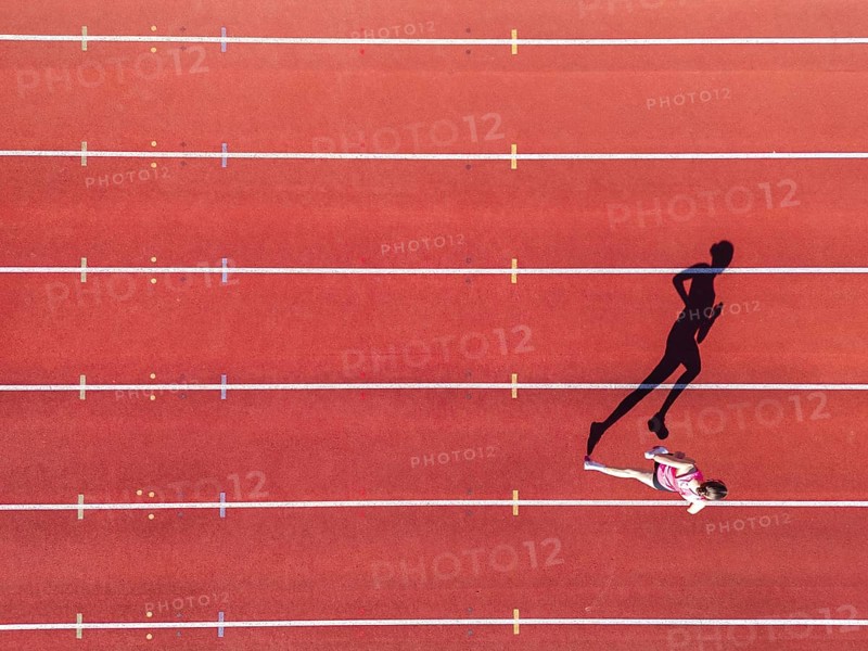 Aerial view of a female individual (runner) on a red athletics track (tartan track) with shadow as a silhouette