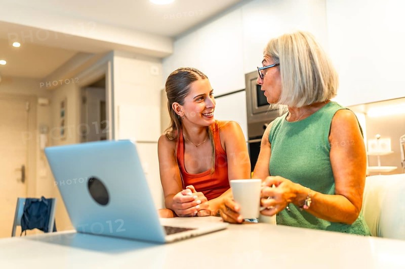 Mother and daughter chatting and smiling while enjoying coffee and browsing a laptop together in a cozy kitchen setting