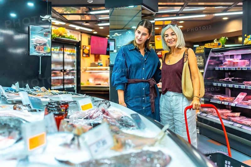 Two women exploring a supermarket's seafood section, examining fresh fish and other products. They appear engaged and interested in their choices