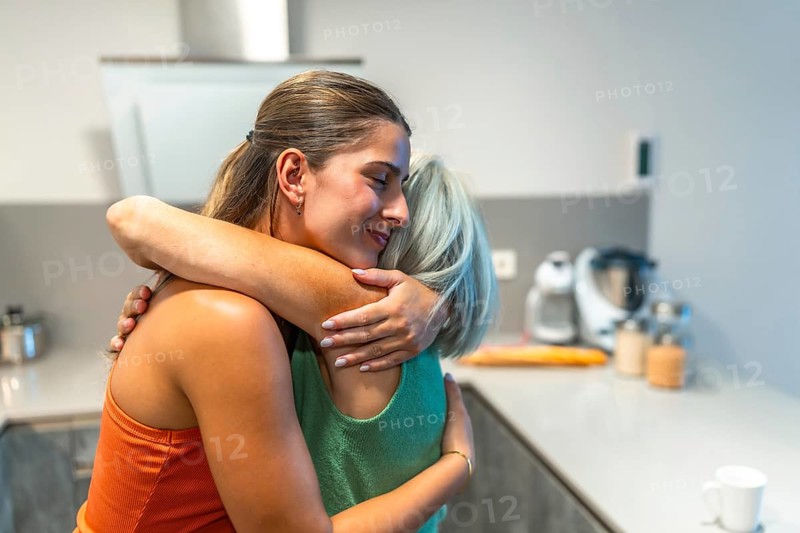 Happy adult daughter is embracing her senior mother in a kitchen, showing love, care, and support