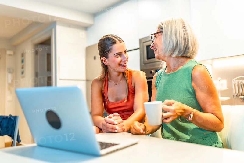Two women sitting at a kitchen counter, engaging in conversation while using a laptop, one enjoying a warm coffee mug, sharing moments together
