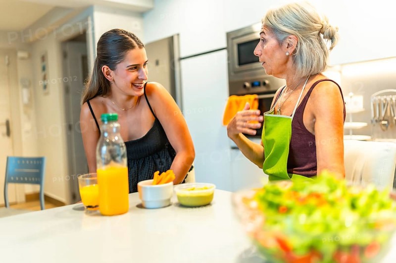 Senior woman wearing apron explaining recipe to her young adult daughter while preparing a healthy meal together in a modern kitchen