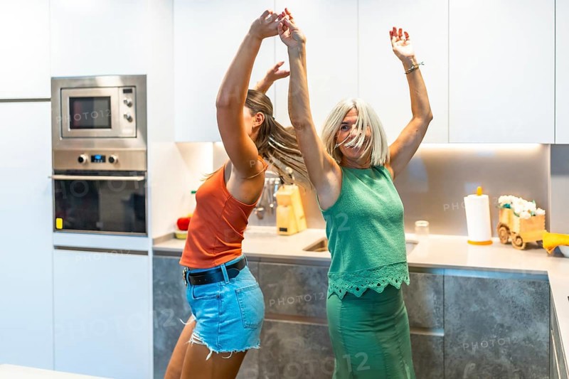 Two women enjoy a lively dance in a contemporary kitchen, celebrating happiness and friendship with joyful expressions and energetic movements