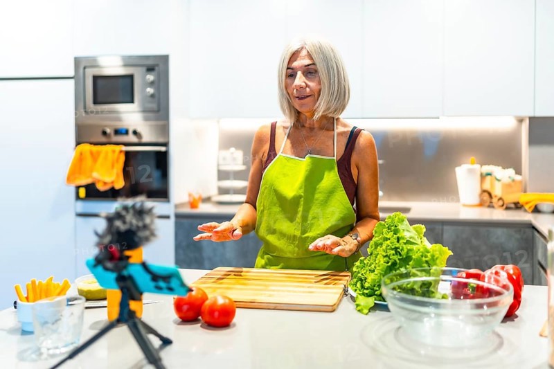 Mature woman in a green apron records a cooking tutorial in a modern kitchen, surrounded by fresh vegetables and kitchen gadgets