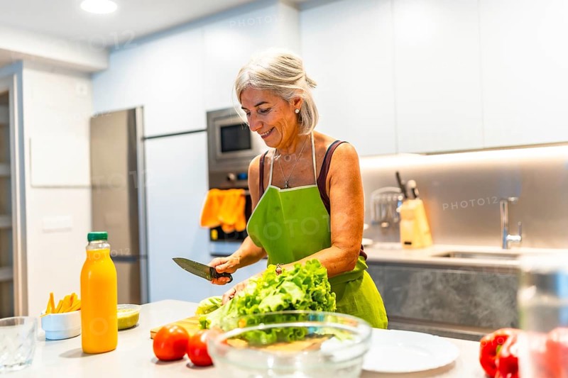 Happy mature woman wearing an apron, preparing a fresh vegetable salad in a modern kitchen filled with vibrant colors and healthy ingredients