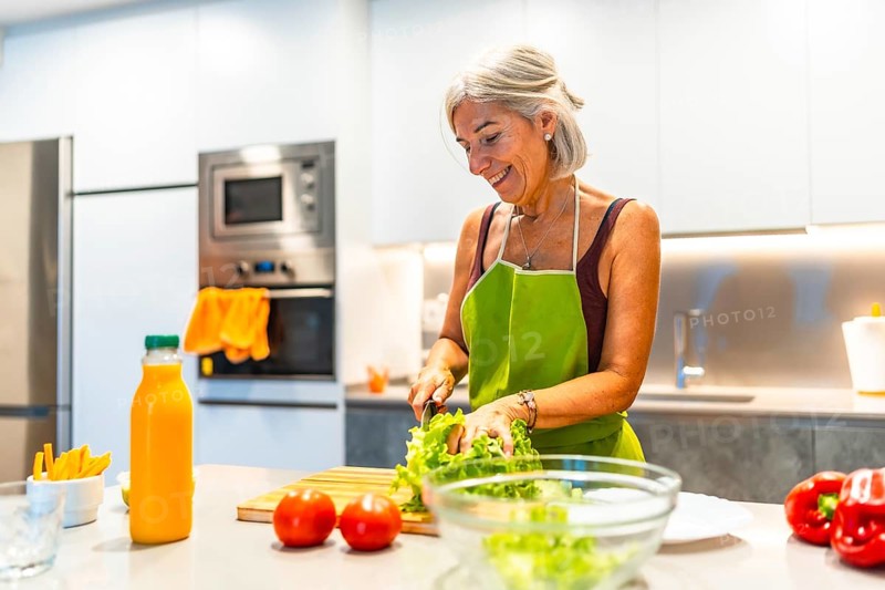 Happy mature woman wearing apron cutting fresh lettuce, preparing healthy meal with organic vegetables in her modern kitchen