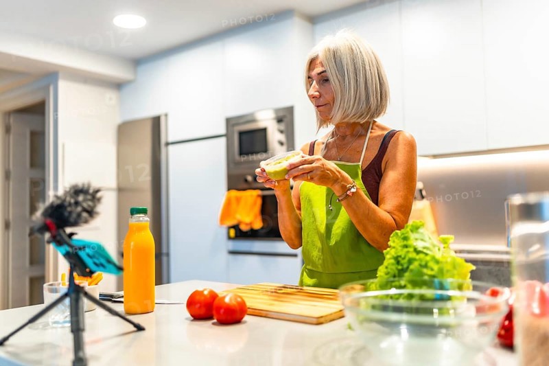 Elderly woman wearing an apron preparing healthy food in her modern kitchen and recording a video recipe with her smartphone