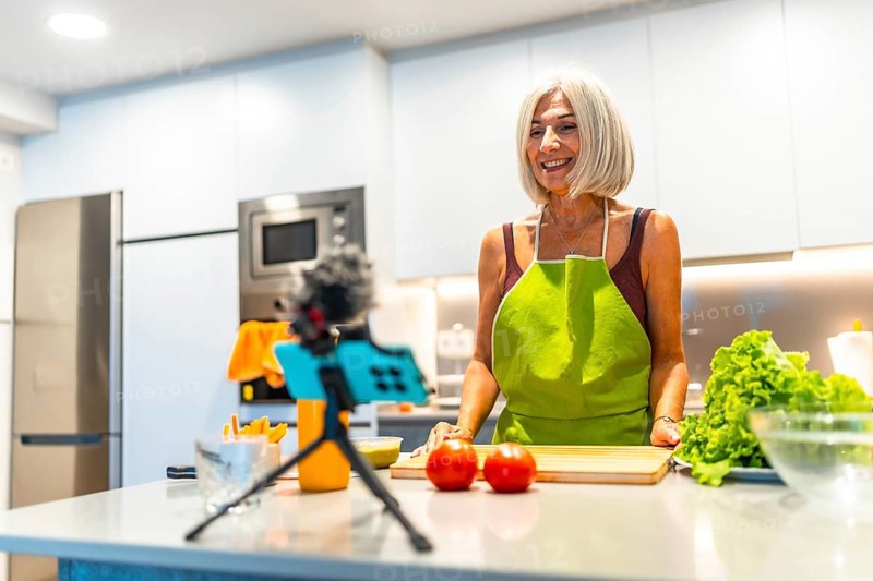 Smiling mature woman wearing apron preparing recipe for her cooking vlog, recording video with smartphone and microphone in kitchen